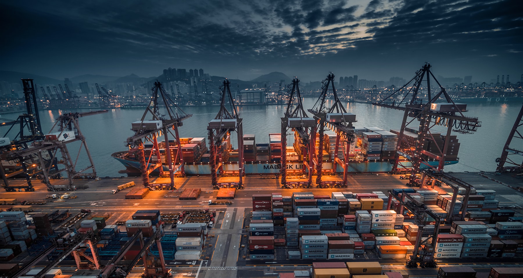 A container ship being worked by gantry cranes at a terminal in the late afternoon.